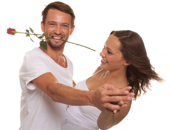 Romantic couple ballroom dancing with the man grinning as he holds a single long stemmed red rose between his teeth to celebrate Valentines day, isolated on white
